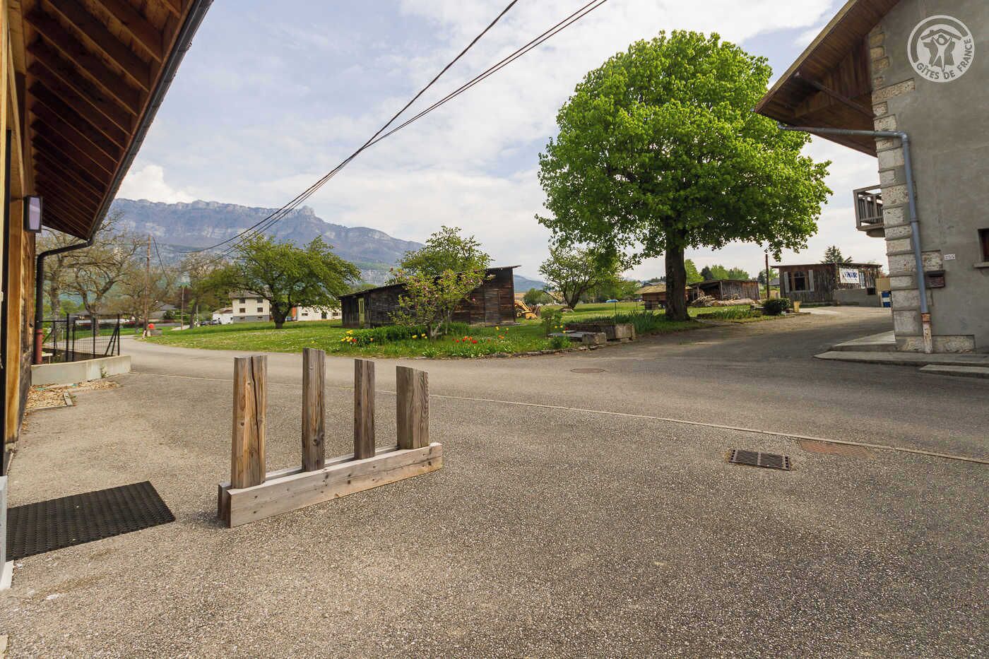 Une rue de campagne avec un bâtiment en bois sur la gauche, un grand arbre au centre et une montagne en arrière-plan.