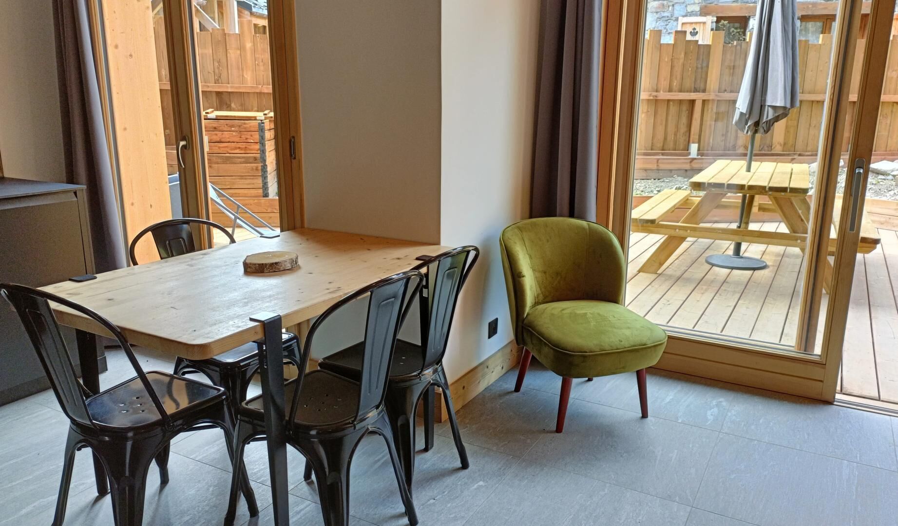 A dining area with a wooden table and black chairs, a green armchair, and a view of an outdoor patio with wooden furniture.