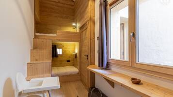 A compact wooden cabin with a small bed, a window with curtains, a wooden bench, and a white chair.
