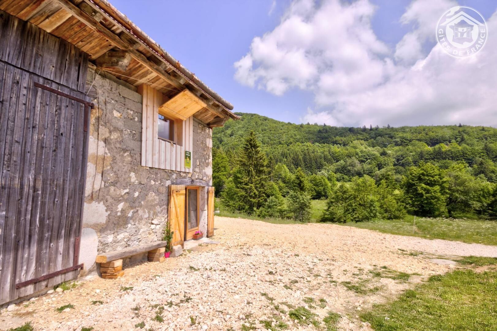 A rustic stone building with a wooden balcony, surrounded by a gravel driveway and lush greenery.