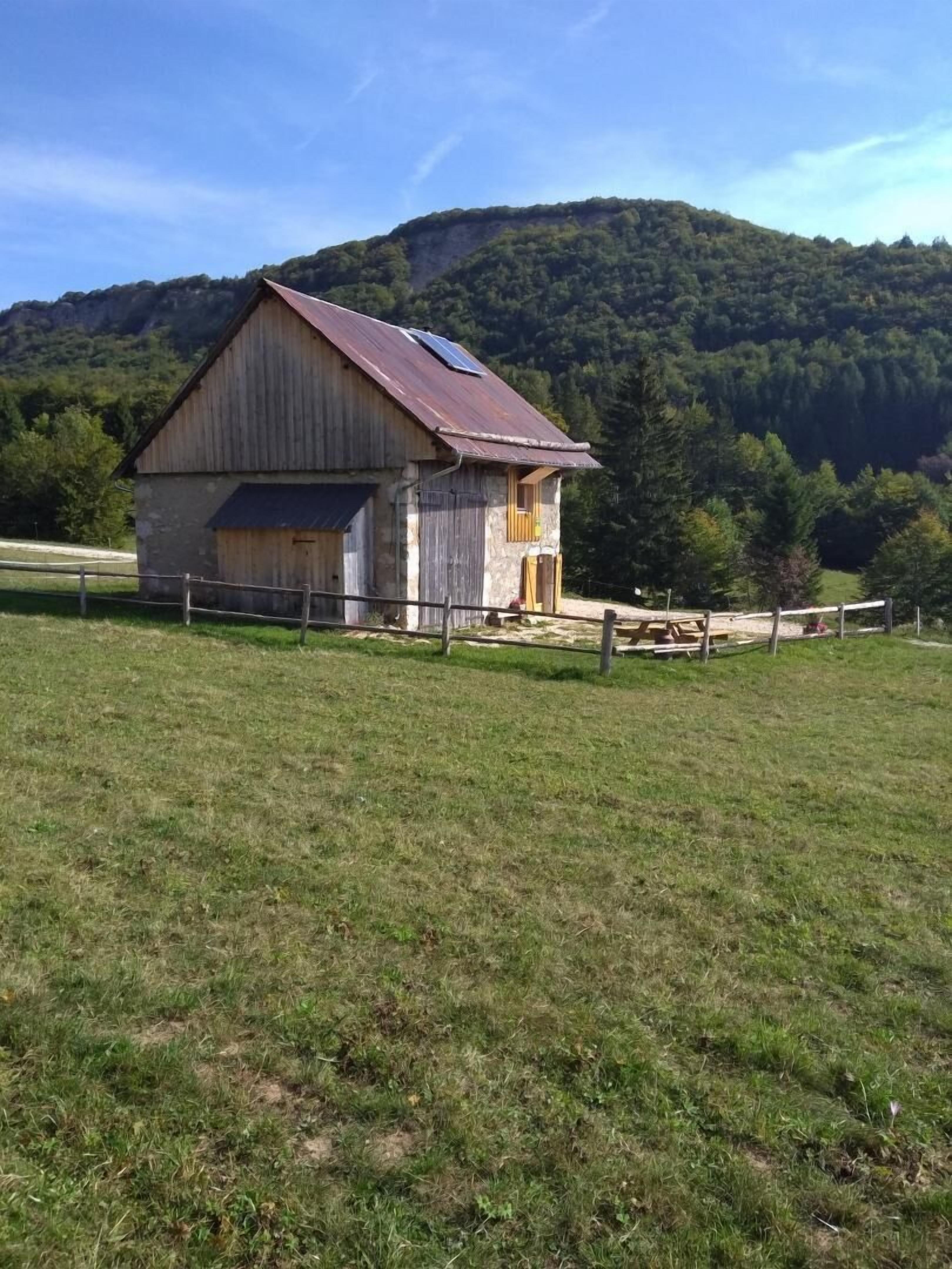Une grange rustique, dotée d’un toit en pente et d’une petite dépendance attenante, entourée d’un pré clôturé avec une montagne en arrière-plan.