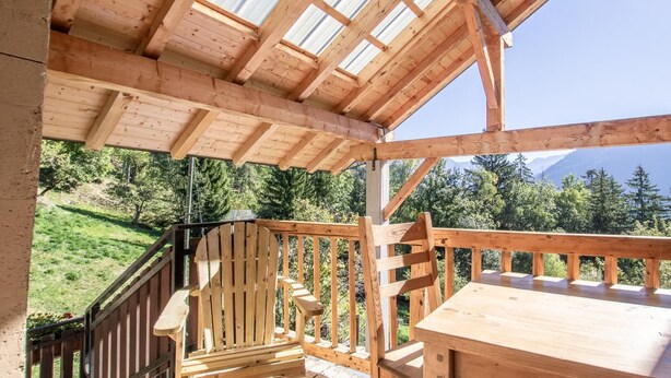 Un patio in legno con tavolo e sedie, una ringhiera di legno e vista su un paesaggio verde con alberi e montagne sullo sfondo.