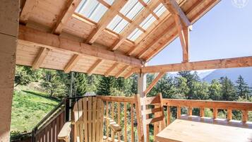 Une terrasse en bois avec une table et des chaises, une rambarde en bois et une vue sur un paysage verdoyant, avec des arbres et des montagnes en arrière-plan.