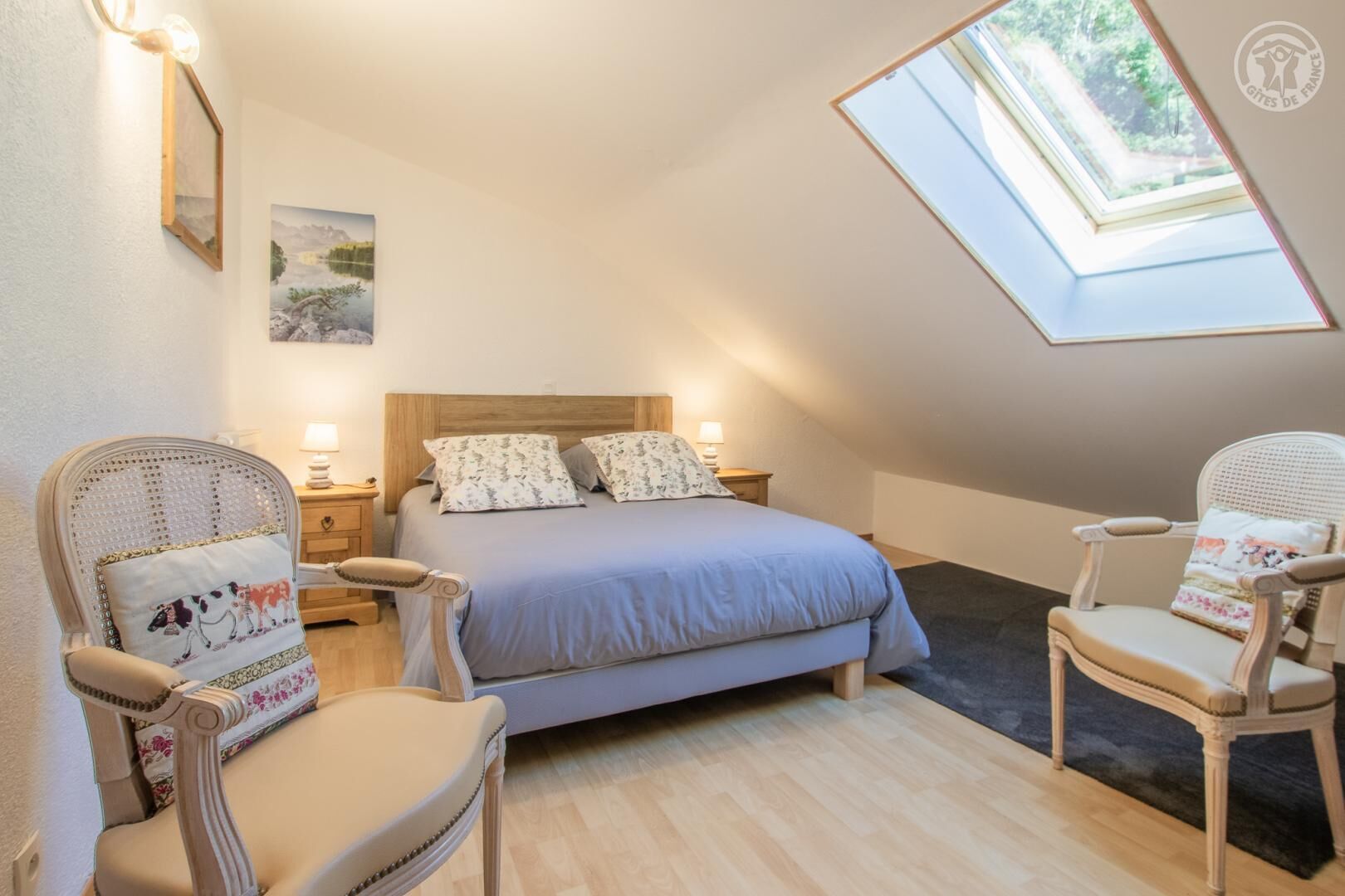 Een slaapkamer met een schuine kap, dakraam, een bed met bloemenkussens, twee nachtkastjes met lampen, en twee rieten stoelen.