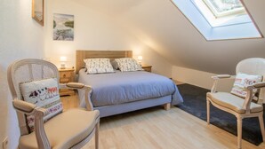 Una camera da letto con soffitto spiovente, un lucernario, un letto con cuscini a motivi floreali, due comodini con lampade e due poltrone in vimini.