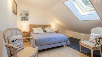 Een slaapkamer met een schuine kap, dakraam, een bed met bloemenkussens, twee nachtkastjes met lampen, en twee rieten stoelen.
