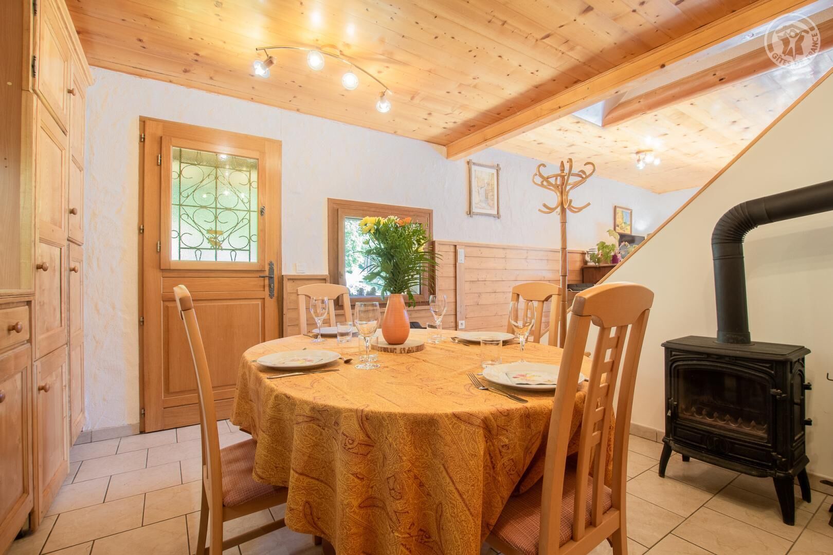 Une salle à manger avec une table ronde dressée pour quatre personnes, des chaises en bois, un vase rempli de fleurs et un poêle à bois noir.