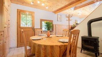 Une salle à manger avec une table ronde dressée pour quatre personnes, des chaises en bois, un vase rempli de fleurs et un poêle à bois noir.