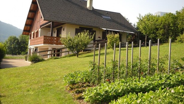 A two-story house with a sloped roof, wooden balcony, and a fenced garden area.