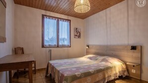 A bedroom with a wooden ceiling, a bed with a patterned bedspread, a window with curtains, a radiator, and a wooden nightstand.