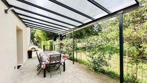 A covered patio area with a table and chairs, surrounded by greenery and a car in the background. - LES LILAS - LA BIOLLE (LA BIOLLE)