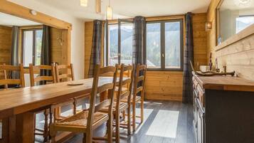 Un coin repas en bois avec une table et des chaises, de grandes fenêtres avec des rideaux et un comptoir de cuisine avec un klaxon.