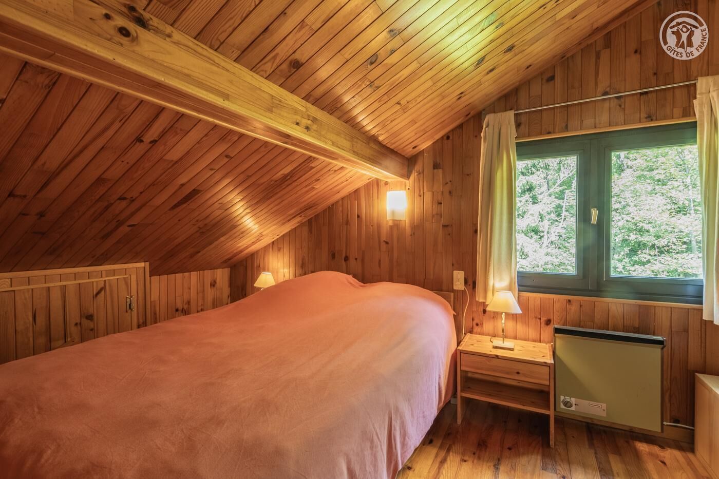 Une chambre confortable située dans les combles, avec un plafond en bois, un lit, deux lampes de chevet, une fenêtre avec des rideaux et un radiateur.