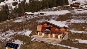 A wooden chalet with a balcony, surrounded by snow-covered hills and trees.