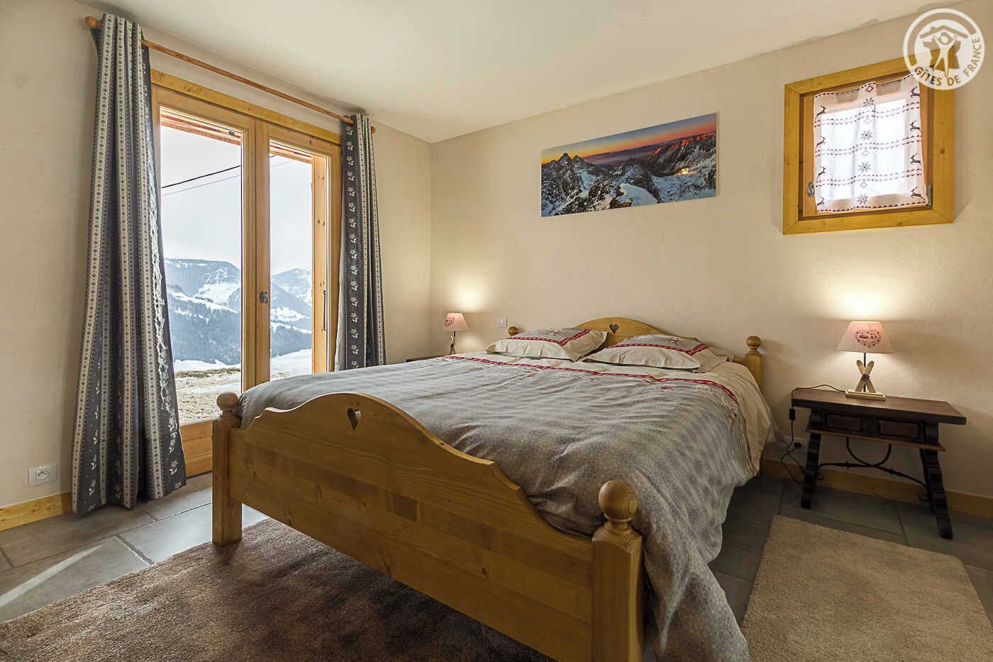 Ein Schlafzimmer mit einem Holzbett, Blick auf Berge durch große Fenster, einem Gemälde an der Wand und einer Nachttischlampe.