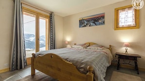 A bedroom with a wooden bed, a view of mountains through large windows, a painting on the wall, and a bedside lamp. - CHALET FIASSE - HAUTELUCE LES SAISIES (HAUTELUCE LES SAISIES)