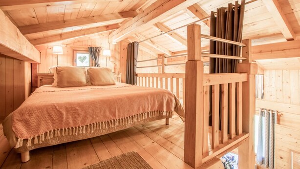 A wooden cabin bedroom with a bed, a wooden railing, and a window with curtains.