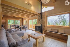 A cozy living room with a fireplace, a dining area, and large windows offering a view of the outdoors.