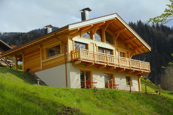 A two-story wooden chalet with a balcony, situated on a grassy hillside with forested mountains in the background.