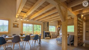 Una habitación amplia con vigas de madera, un comedor con sillas y mesa, una sala de estar con sofá y sillones, y ventanas grandes que ofrecen vistas al exterior.