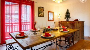 Ein Essbereich mit Holztisch, gedeckten Plätzen für vier Personen, roten Servietten und Kerzen. An der Wand befinden sich ein kleiner Weihnachtsbaum und ein gerahmtes Bild.