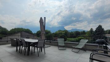 A rooftop terrace with a table and chairs, a barbecue grill, and a view of mountains and trees.