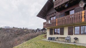 A chalet-style building with a balcony, outdoor seating, and a view of a mountainous landscape. - CHALET DES CIMES - NOTRE DAME DU PRE (NOTRE DAME DU PRE)