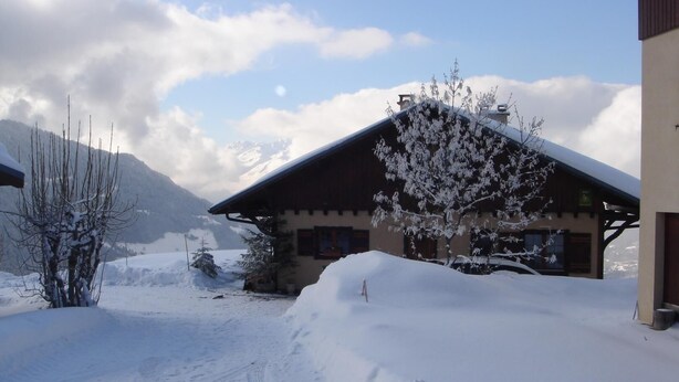 A snow-covered building with a sloped roof, a snow-covered tree in front, and a mountain in the background. - CHALET DES CIMES - NOTRE DAME DU PRE (NOTRE DAME DU PRE)