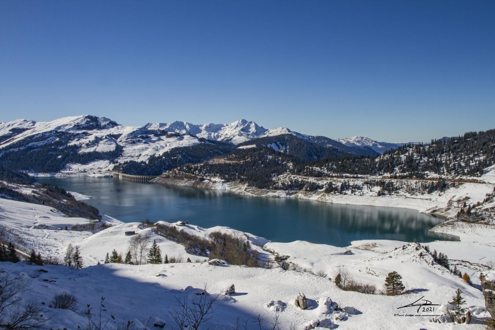 A panoramic view of a snow-covered landscape with a lake, mountains, and a bridge.