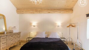 A bedroom with a wooden ceiling, a bed with white and black bedding, bedside lamps, a dresser with a mirror, and a hanging rack. - LE MAZOT - VILLAROGER (VILLAROGER)