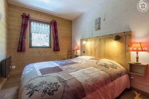 Une chambre chaleureuse avec une tête de lit en bois, un lit recouvert d’une courtepointe à motifs floraux, deux lampes de chevet et une fenêtre avec des rideaux.