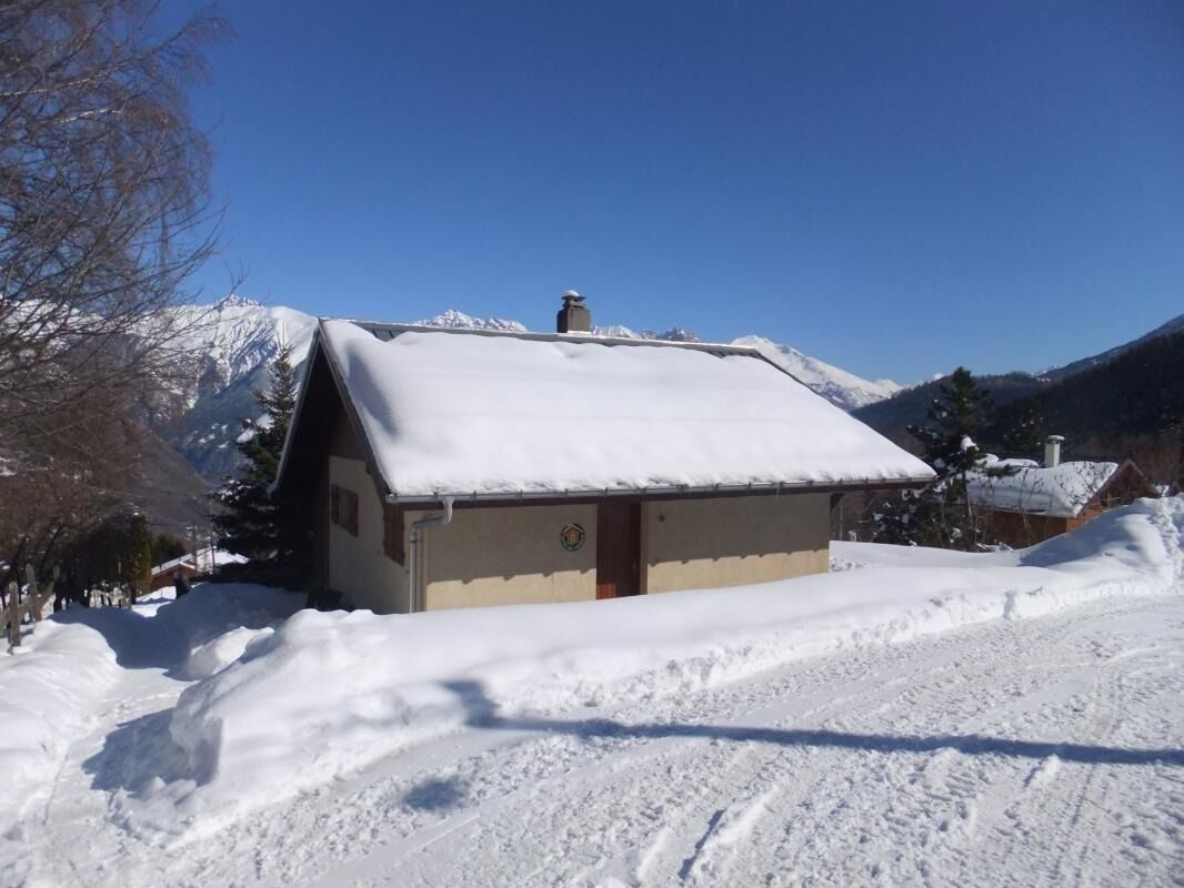 A small, snow-covered building with a sloped roof, surrounded by snow and mountains in the background.