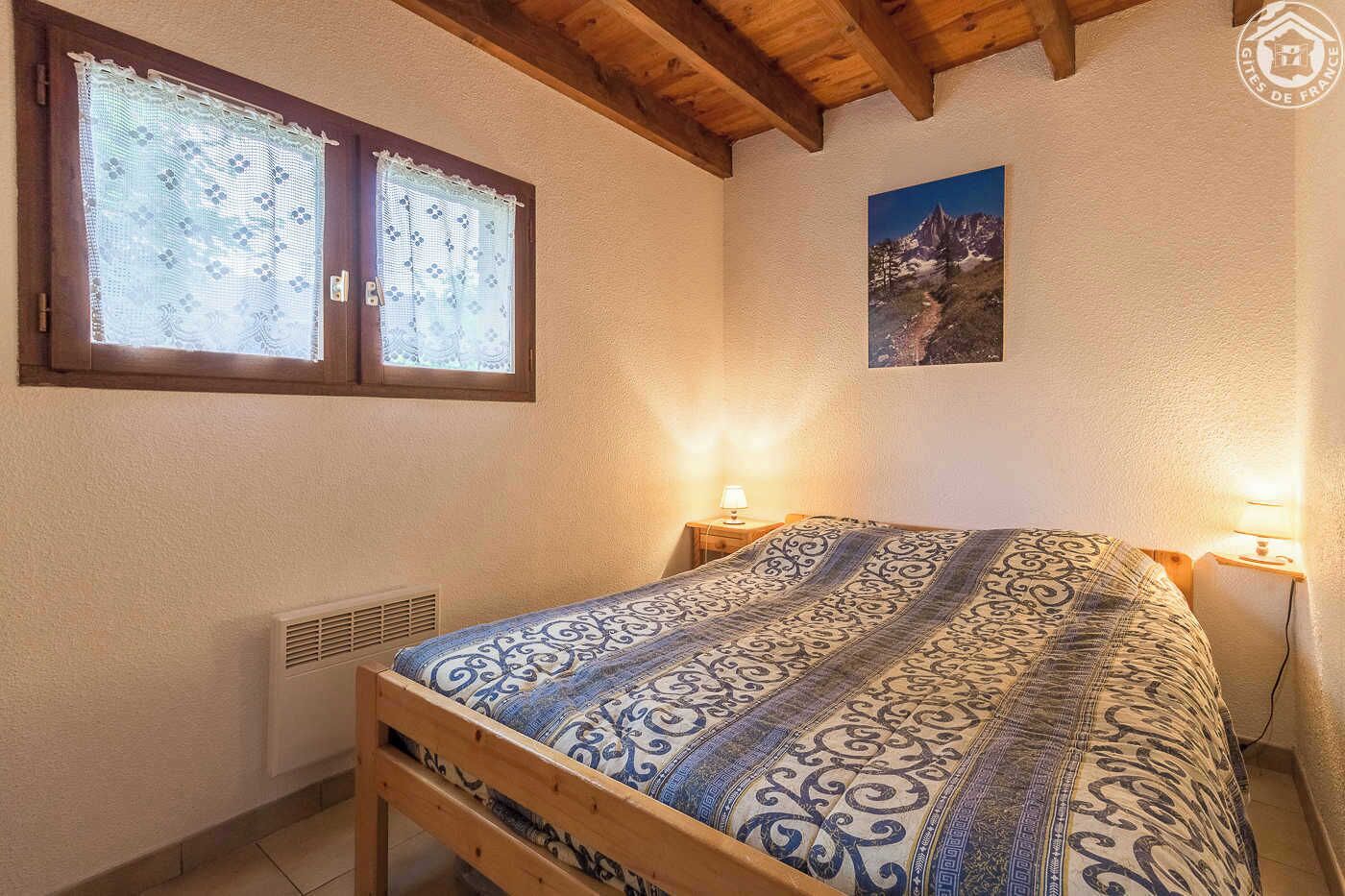 Een slaapkamer met een houten bed, twee bedlampen, een raam met kantgordijnen en een schilderij van een berglandschap aan de muur.