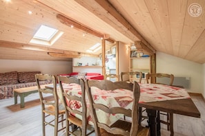 Um quarto de sótão acolhedor com teto de madeira, uma mesa de jantar com cadeiras, um sofá e uma cozinha com prateleiras.