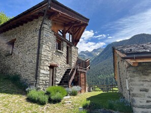 A stone building with a wooden staircase leading to a balcony, surrounded by greenery and mountains in the background.