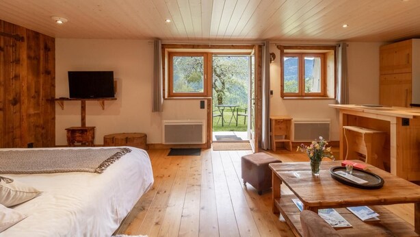Ein Zimmer aus Holz mit einem Bett, einem Fernseher, einem Tisch mit einer Vase und Blick ins Freie.
