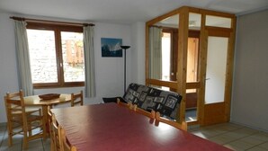 Un coin repas avec une table rouge, des chaises en bois, une petite table ronde avec un panier, et une vue sur un bâtiment en pierre par la fenêtre.