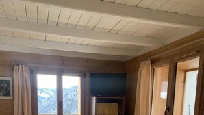 En stuga med träpanel på insidan, inredd med en soffa, två gungstolar, en bokhylla och en utsikt över bergen genom ett fönster.