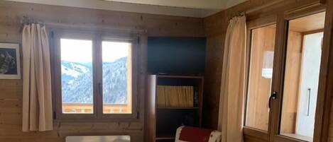 Ein Holzblockhaus-Innenraum mit einer Couch, zwei Schaukelstühlen, einem Bücherregal und Blick auf Berge durch ein Fenster.