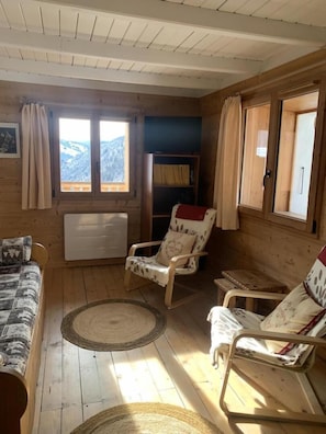 A wooden cabin interior with a sofa, two rocking chairs, a bookshelf, and a view of mountains through a window.