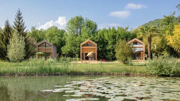 Drei Holzhütten mit offenen Vorderseiten, gelegen an einem Teich mit Seerosen, umgeben von üppiger Vegetation und Bäumen.