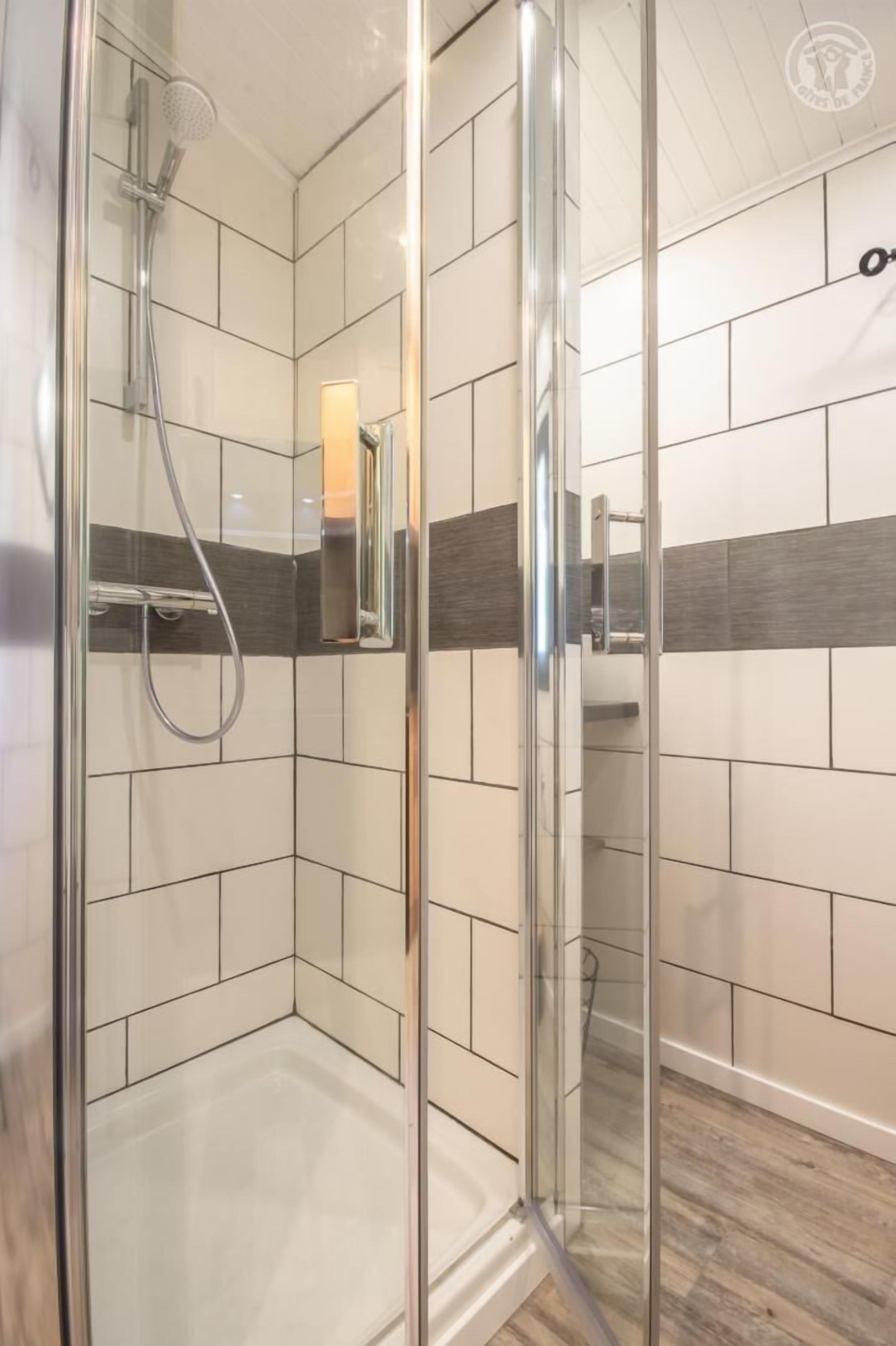 A modern shower with a glass door, white and grey tiles, and a wooden floor.