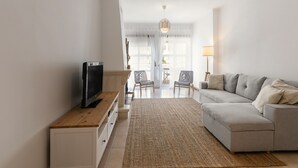 Ett modernt vardagsrum med en platt-TV på ett träställ, en grå soffa med kuddar, en vävd matta och en matplats med stolar och ett bord i bakgrunden.