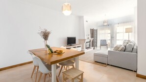 Ett modernt vardagsrum med ett matbord i trä, vita stolar, en grå soffa och en tv.