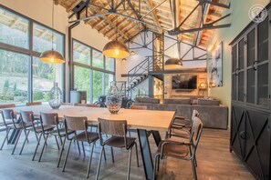 Een ruime eethoek met een lange houten tafel en moderne stoelen, grote ramen met uitzicht op de omgeving, en een hoge plafond met zichtbare balken.