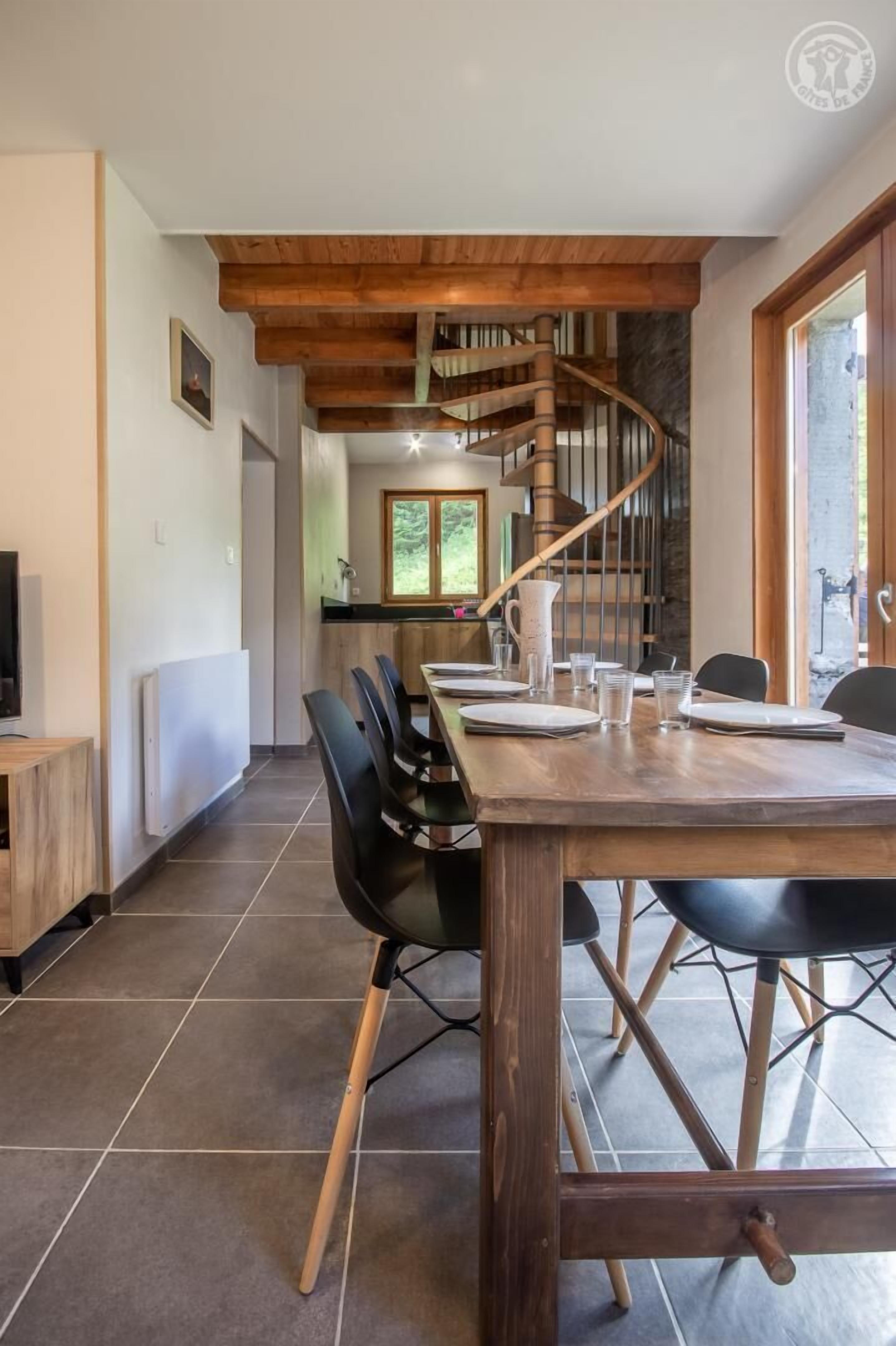 Un coin repas avec une table en bois dressée pour quatre personnes, des chaises noires et un escalier en colimaçon en arrière-plan.