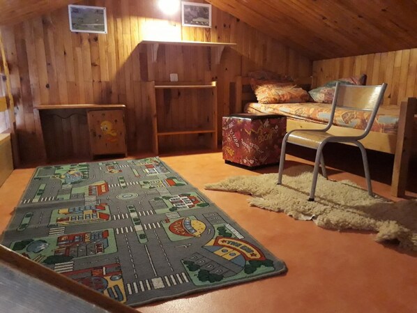 A small attic bedroom with wooden walls, a bed with patterned bedding, a chair, a small table, and a rug with a cityscape design.