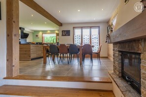 A dining area with a wooden table and chairs, a kitchen in the background, and a fireplace.