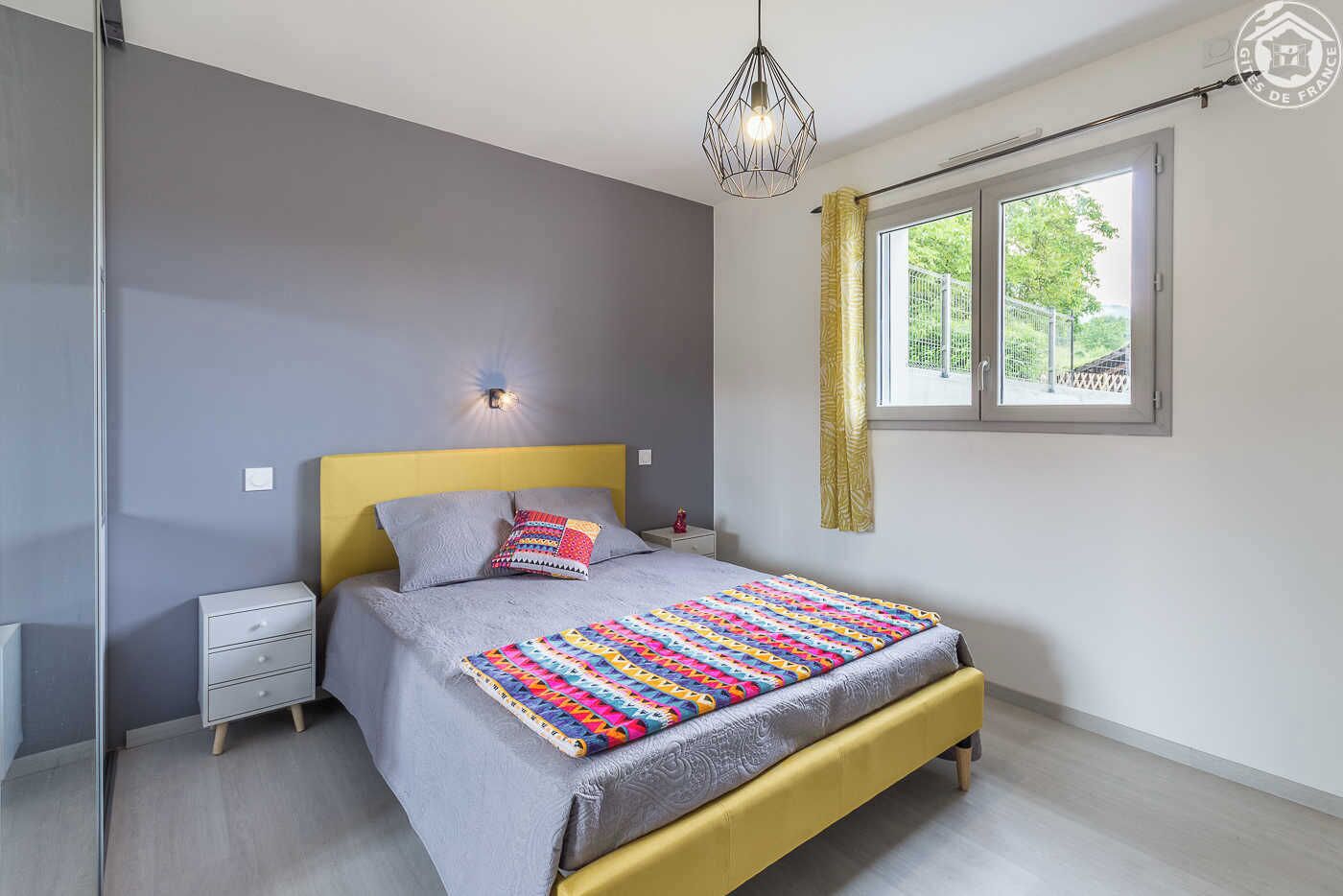 Quarto moderno com cama, mesas de cabeceira, janela com cortinas e luminária suspensa.