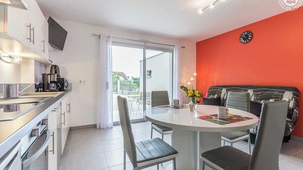 A modern kitchen with a built-in oven, a dining area with a round table and four chairs, a red accent wall, and a large window with a view of the outdoors.